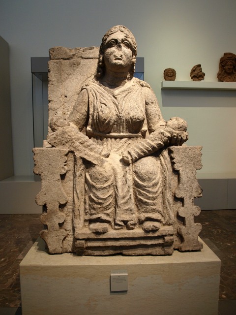 Escultura en piedra de mujer sentada, hierática, que mira de frente al espectador. Sobre su regazo, fajados y colocados uno frente al otro, dos niños de corta edad.
