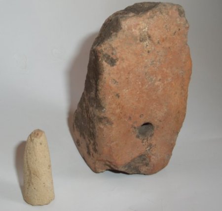 Dos fragmentos de cerámica. Uno es una pesa de telar muy dañada, de color rojizo. El otro tiene el tamaño de un dedo meñique y es de barro claro.