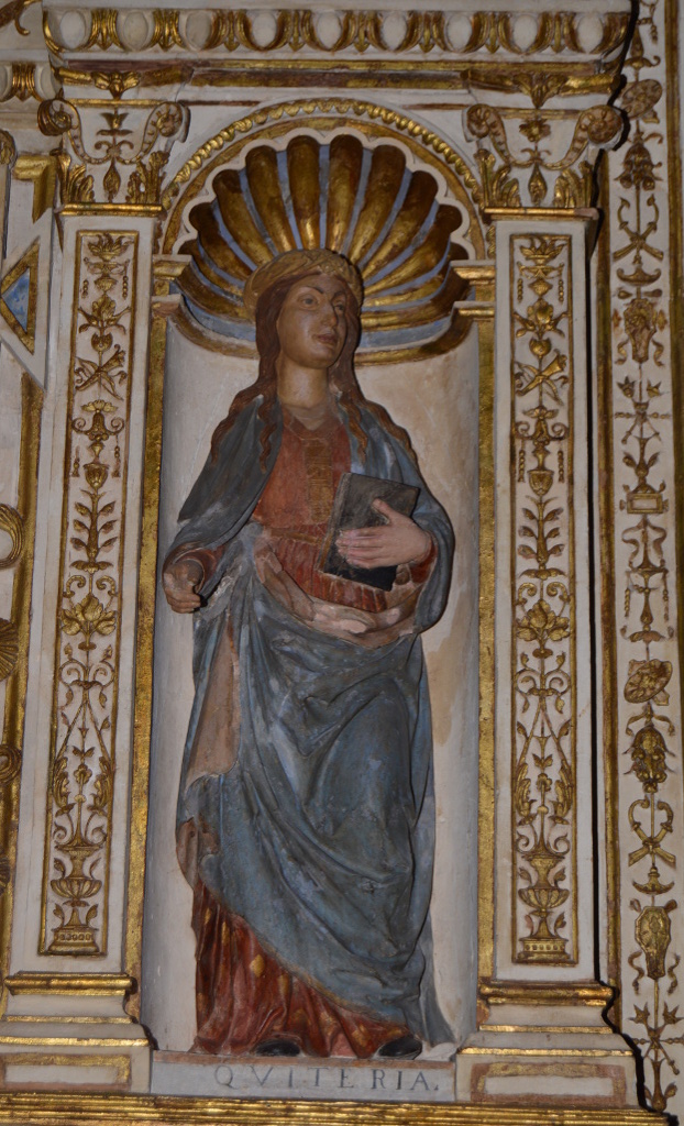 Escultura de Santa Quiteria representada erguida, vestida de azul, con un libro en la mano izquierda. En la mano derecha debía sostener algo, que se ha perdido